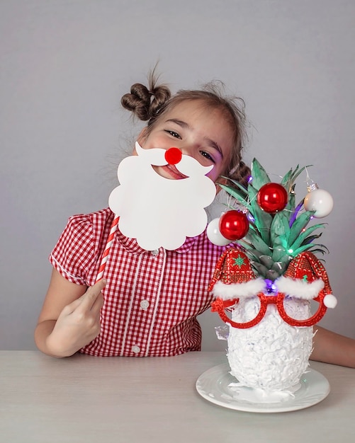 Fröhliches süßes kleines Mädchen in der Nähe des Weihnachtsmann-Gesichts aus Ananas