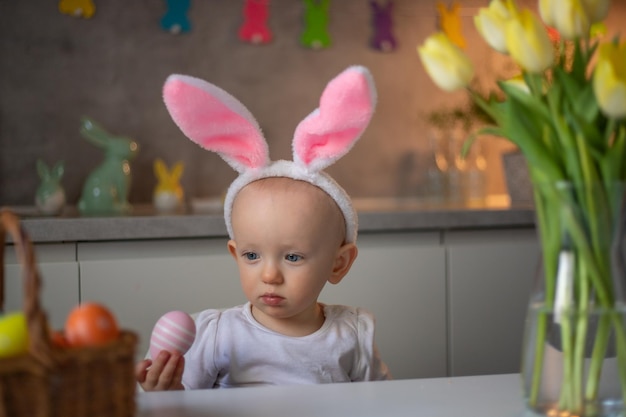 Fröhliches süßes kleines Mädchen mit Hasenohren am Ostertag, das am Tisch in der Küche sitzt