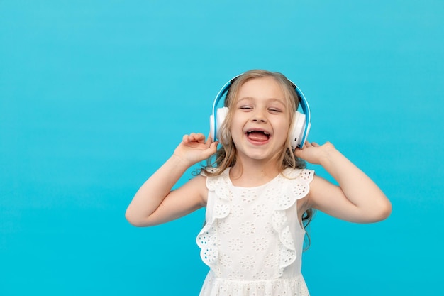 Fröhliches süßes kleines Mädchen mit Kopfhörern, das Musik in einem weißen Baumwollkleid auf blauem Hintergrund im Studio hört und lächelt, Platz für Text