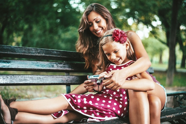 Fröhliches süßes kleines Mädchen und ihre Mutter posieren für ein Selbstporträt auf einer Bank im Park.