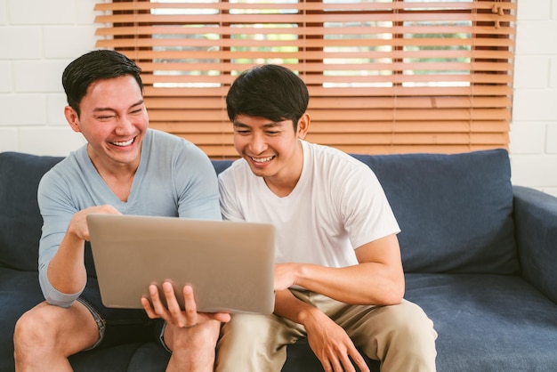 Fröhliches tausendjähriges asiatisches schwules Paar, das auf dem Boden sitzt und sich auf ein Sofa lehnt, während es sich im Wohnzimmer zu Hause Inhalte auf einem Laptop ansieht LGBT-Multibeziehung Konzept für schwule Paare