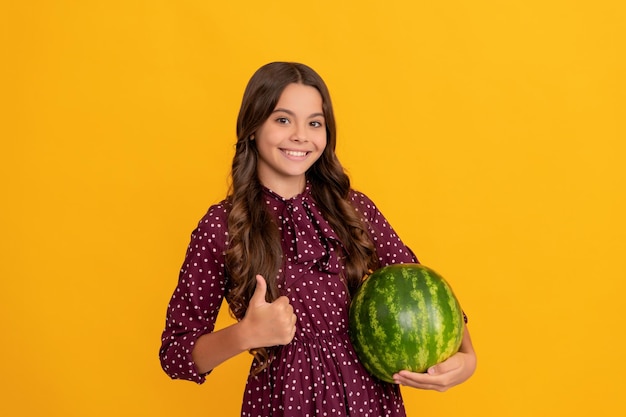Fröhliches Teenager-Mädchen hält frische reife Wassermelonenfrüchte, die den Daumen nach oben im Sommer zeigen