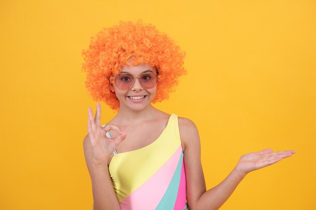 Fröhliches Teenager-Mädchen mit Sonnenbrille auf gelbem Hintergrund, das Produkt mit ok-Kopie-Werbung präsentiert