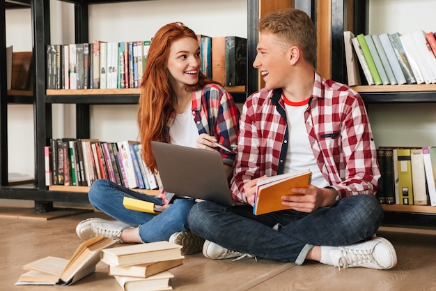 Fröhliches Teenagerpaar, das auf einem Boden am Bücherregal sitzt