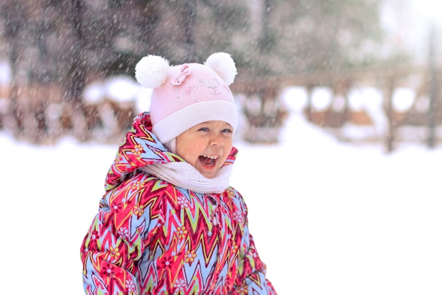 Fröhliches und lächelndes kleines Mädchen, das im verschneiten Winterpark auf Schnee lacht