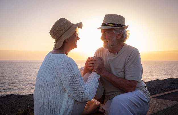 Fröhliches und romantisches kaukasisches Paar, das die Hände in den Händen hält, die draußen am Meer bei Sonnenuntergang sitzen und in die Augen alter Senioren oder Rentner schauen, die gemeinsam Urlaub machen