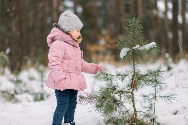 Fröhliches Winterkonzept. Ein kleines Mädchen in einer rosa Jacke und einer grauen Mütze spielt mit einem Kiefernzweig und lächelt. Glückliches Mädchen im verschneiten Winterwald. Platz kopieren.