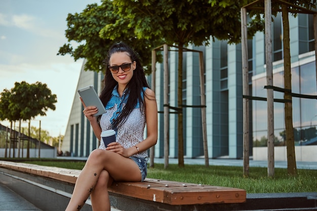 Fröhliches, wunderschönes brünettes Mädchen mit Tattoo auf ihrem Bein, das trendige Kleidung und Sonnenbrille trägt, hält einen Kaffee zum Mitnehmen und ein digitales Tablet, während sie auf einer Bank gegen einen Wolkenkratzer sitzt und einen Kam betrachtet