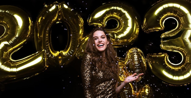 Fröhliches wunderschönes Mädchen im stilvollen sexy Partykleid mit goldenen 2023-Ballons, die sich auf der Silvesterparty amüsieren