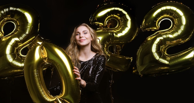 Fröhliches wunderschönes Mädchen in stilvollem sexy Partykleid mit goldenen Luftballons 2022, das Spaß bei der Weihnachts- oder Silvesterparty hat.