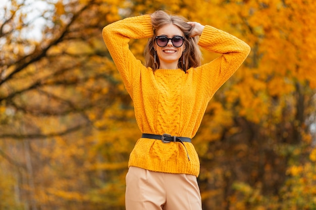 Fröhliches, zufriedenes junges Mädchen mit einem Lächeln in modischer Sonnenschutzbrille in einem gestrickten gelben Pullover geht auf dem Land mit orangefarbenen Herbstblättern
