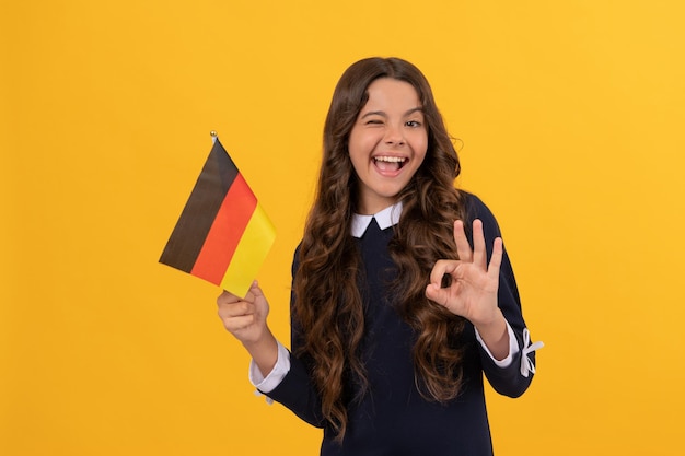 Fröhliches zwinkerndes Kind hält den gelben Hintergrund der deutschen Flagge, der eine ok Geste zeigt, Bildung im Ausland.