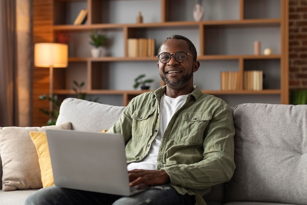 Froh, dass ein afrikanisch-amerikanischer Mann mittleren Alters mit Brille einen Videoanruf auf einem Laptop hat, der im Wohnzimmer im Internet surft