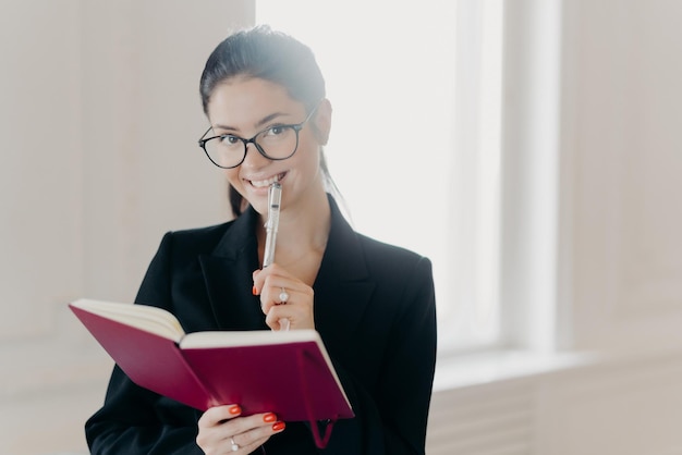 Froh, erfolgreicher Budgetanalyst notiert etwas Text in Notizbuch, lächelt glücklich, hält Stift, trägt elegantes schwarzes, formelles Outfit, transparente Brille, posiert in geräumigem Raum in der Nähe des Fensters, macht Checkliste