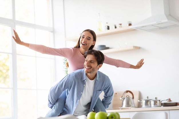 Froh, glücklicher junger Ehemann trägt seine Frau auf dem Rücken und hat gemeinsam Spaß im Kücheninternet