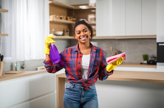 Froh, tausendjährige schwarze Frau in Handschuhen mit Sprühlappen hat Spaß und genießt die Reinigung mit professionellen Reinigungsmitteln