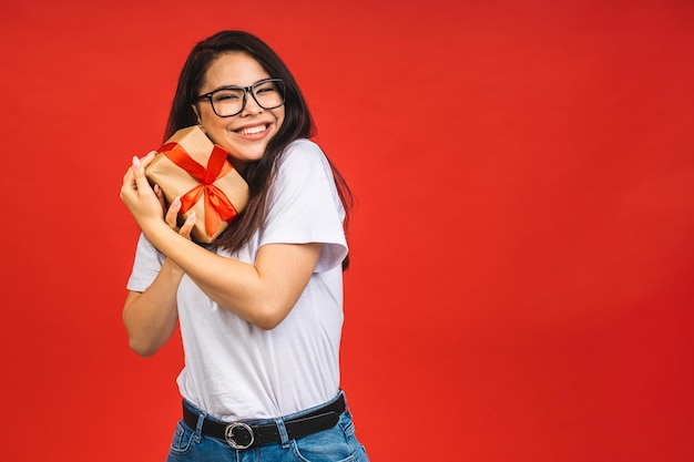 Frohe Feiertage Studioporträt der lächelnden jungen schönen Brunettefrau hält die Geschenkbox, die über rotem Hintergrund lokalisiert wird