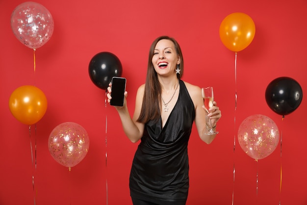 Frohe junge Frau im schwarzen Kleid mit Glas Champagner, Handy mit leerem schwarzen leeren Bildschirm auf leuchtend roten Hintergrundluftballons. Frohes neues Jahr, Geburtstagsmodell Urlaub Party Konzept.