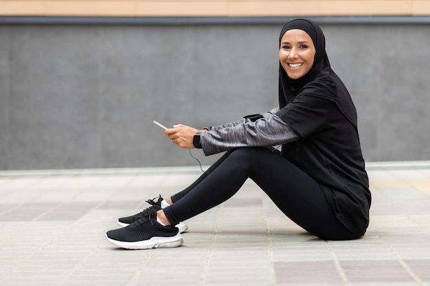 Frohe junge islamische Sportlerin in Hijab-Sportbekleidung mit Smartphone sitzt auf dem Boden an grauer Wand