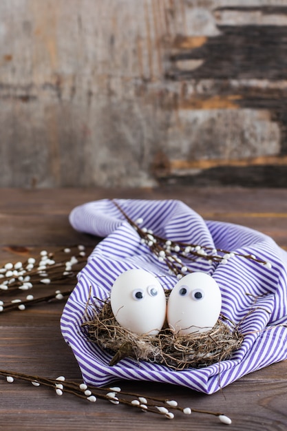 Frohe Ostern. Ein paar ungefärbte Eier mit Augen in einem Vogelnest und Weidenzweigen auf einem Holztisch.