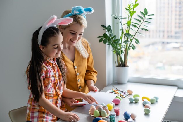 Frohe Ostern. Eine Mutter und ihre Tochter bemalen Ostereier. Glückliche Familie bereitet sich auf Ostern vor. Nettes kleines Mädchen mit Hasenohren am Ostertag.