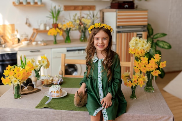 Frohe Ostern fröhliches schönes Mädchen in einem grünen Kleid mit Blumenkranz spielt mit einem Kaninchen zu Hause in der Küche