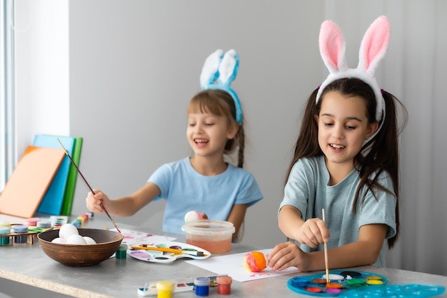 Frohe Ostern. Zwei Schwestern, die Ostereier bemalen. Glückliche Familienkinder, die sich auf Ostern vorbereiten. Süßes kleines Mädchen mit Hasenohren am Ostertag.