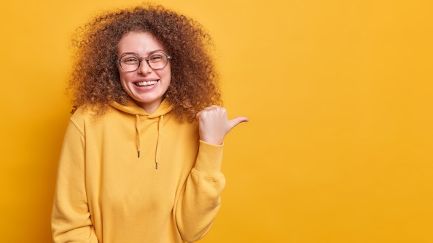 Frohe positive lockige Frau in Brillen lächelt aufrichtig wirbt für das Produkt hat gute Laune und zeigt Kopienraumpunkte mit dem Daumen, der in einem Sweatshirt isoliert über der gelben Wand gekleidet ist