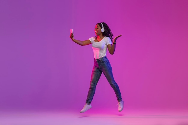 Frohe schwarze Frau im Neonlicht, das mit Smartphone in den Händen springt