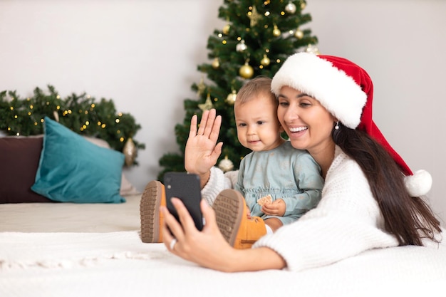 Frohe Weihnachten Eine schöne Mutter und eine kleine Tochter rufen ihre Verwandten per Videolink an oder machen ein Selfie auf einem Smartphone Gemütlich eingerichtetes Schlafzimmer Lifestyle