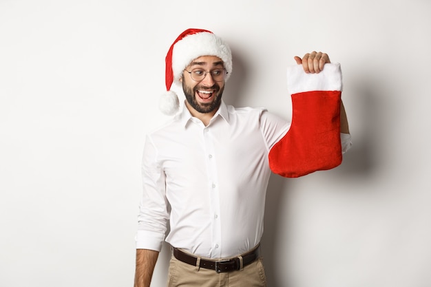 Frohe Weihnachten, Feiertagskonzept. Glücklicher erwachsener Mann erhalten Geschenke in der Weihnachtssocke, die aufgeregt schaut und Weihnachtsmütze, weißen Hintergrund trägt.