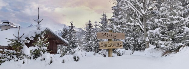 Frohe Weihnachten geschrieben auf Holzpostschild neben einer Almhütte und einem Wald im Schnee