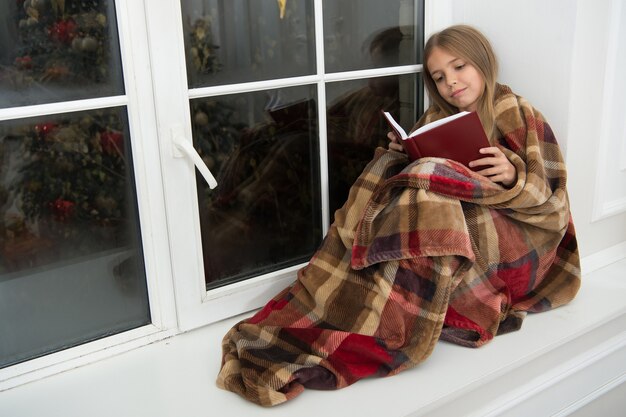 Frohe Weihnachten. Kleines Mädchen genießt es, Weihnachtsgeschichte zu lesen. Kleines Kind las am Heiligabend ein Buch. Kleiner Leser in Plaid gehüllt sitzt auf der Fensterbank. Bilderbuch kinder. Zauberhafte Weihnachtsstimmung.