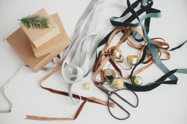 Foto frohe weihnachten nachhaltige feiertage, die stilvolle öko-weihnachtsgeschenke verpacken einfache bastelgeschenkboxen samtbänder weihnachtsglocken tannenzweig und schere auf weißem hintergrund