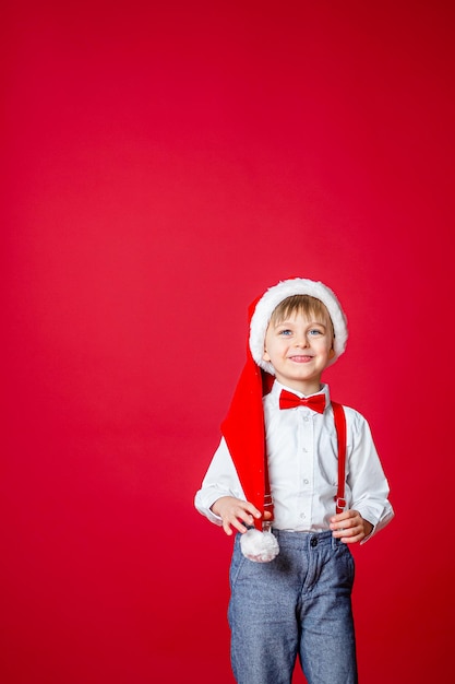 Frohe Weihnachten Netter fröhlicher kleiner Junge im Weihnachtsmann-Hut auf rotem Hintergrund glückliche Kindheit