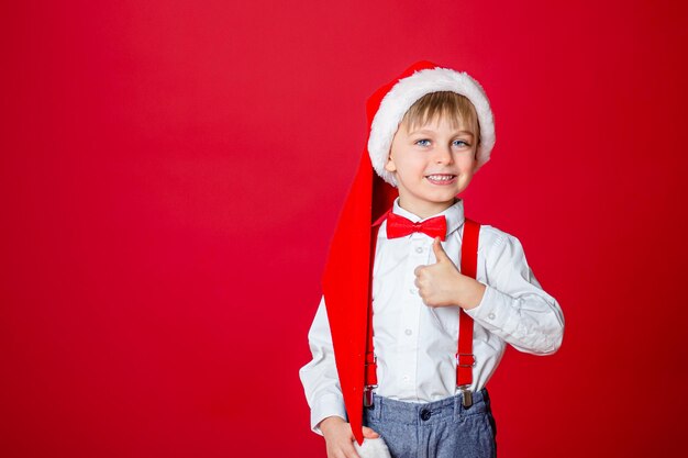 Frohe Weihnachten Netter fröhlicher kleiner Junge im Weihnachtsmann-Hut auf rotem Hintergrund glückliche Kindheit