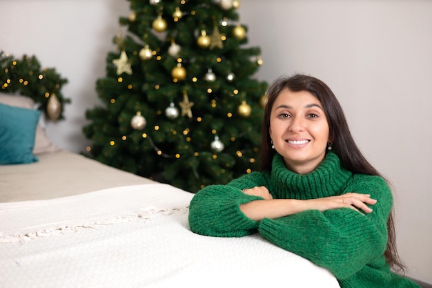 Frohe Weihnachten Porträt einer schönen jungen Frau in einem gemütlichen Schlafzimmer mit einem geschmückten Weihnachtsbaum im Hintergrund Grüner Pullover Lifestyle