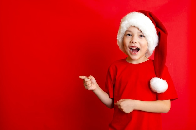 Frohe Weihnachten. Porträt eines netten fröhlichen Jungen in einer Weihnachtsmütze in einem roten T-Shirt auf rotem Grund. Ein Platz für Texte. Foto in hoher Qualität