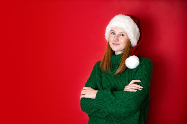 Frohe Weihnachten Porträt eines schönen jungen Mädchens im Teenageralter in einem gemütlichen grünen Strickpullover und Santa39s Hut Der rote Hintergrund ist der Platz für den Text