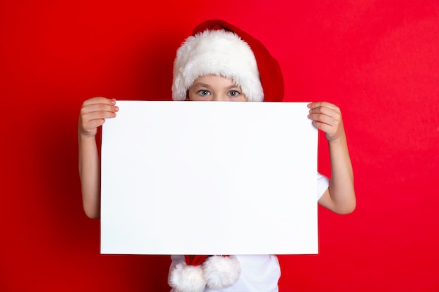 Frohe Weihnachten. Porträt eines süßen Jungen in einer Weihnachtsmütze mit einem weißen Blatt in den Händen auf rotem Grund. Platz für Text. Foto in hoher Qualität