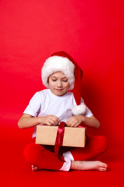 Frohe Weihnachten. Porträt eines süßen Jungen in einer Weihnachtsmütze mit Geschenkboxen auf rotem Grund. Ein Platz für Texte. Foto in hoher Qualität