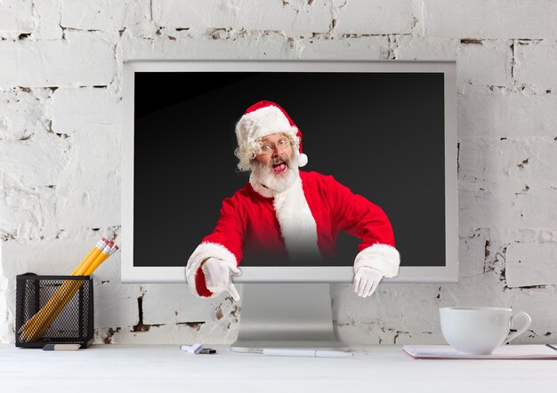 Frohe Weihnachten Santa Claus versteckt im Monitor auf weißem Backsteinmauerhintergrund. Kaukasisches männliches Modell in der Tracht des traditionellen Feiertags. Konzept von Feiertagen, Neujahr, Winterstimmung, Geschenken. Exemplar.