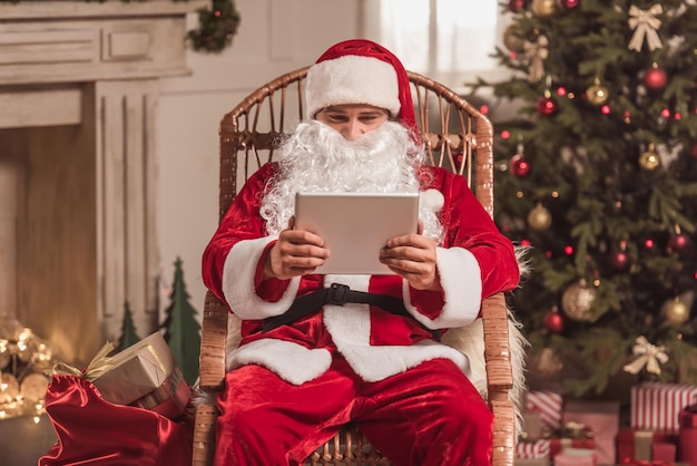 Frohe Weihnachten und ein glückliches Neues Jahr! Der Weihnachtsmann benutzt ein digitales Tablet, während er im Schaukelstuhl neben dem Weihnachtsbaum sitzt