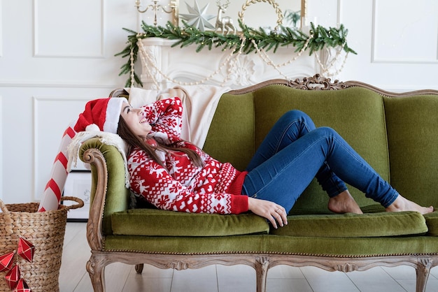 Frohe Weihnachten und ein glückliches Neues Jahr. Junge brünette Frau mit Weihnachtsmütze, die allein auf der grünen Couch in einem für Weihnachten dekorierten Wohnzimmer liegt?