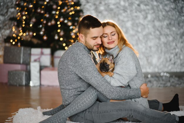 Frohe Weihnachten und ein glückliches Neues Jahr. Junges schönes Paar spielt mit ihrem Hund im festlichen Neujahrswohnzimmer kurz vor Weihnachten.