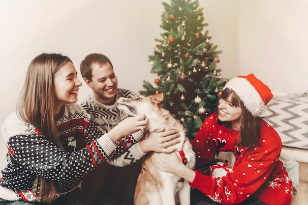 Frohe weihnachten und ein gutes neues jahr konzept stilvolle hipster-familie in festlichen pullovern, die mit süßem hund am weihnachtsbaum spielen und lächeln, beleuchtet frohe feiertage atmosphärische emotionale momente