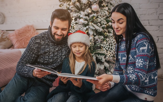 Frohe Weihnachten und schöne Feiertage Fröhliche Mama, Papa und ihr süßes Tochtermädchen tauschen Geschenke aus. Eltern und kleines Kind, die Spaß nahe Weihnachtsbaum drinnen haben. Morgen Weihnachten.