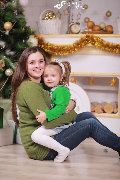 Frohe Weihnachten und schöne Feiertage. Hübsche junge Mutter mit ihrer niedlichen kleinen Tochter nahe Weihnachtsbaum mit Geschenkboxen drinnen.