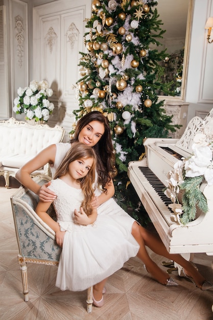 Frohe Weihnachten und schöne Feiertage. Nette Mutter und ihr nettes Tochtermädchen im weißen klassischen weißen Innenklavier und in einem verzierten Weihnachtsbaum. Neujahr