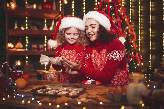 Frohe Weihnachten und schöne Feiertage. Nettes nettes gelocktes kleines Mädchen und ihre ältere Schwester in Sankt-Hüten Weihnachtsplätzchen kochend.